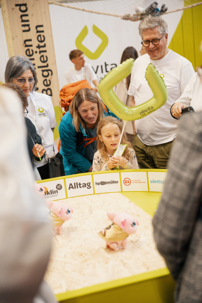 Eine Gruppe von Erwachsenen und Kindern beobachtet bei einer Veranstaltung zwei Spielzeugschweinchen in einem kleinen Gehege. Ein Mädchen hält einen Luftballon in der Hand, und die Menschen wirken engagiert und fröhlich. Im Hintergrund ist ein "avita"-Banner zu sehen.