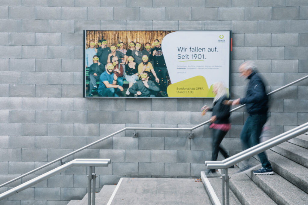 Zwei Personen gehen eine Treppe hinauf und stehen vor einer grauen Backsteinmauer, auf der ein Plakat ein Gruppenfoto und einen deutschen Text zeigt: "Wir fallen auf. Seit 1901". Das Bild ist leicht unscharf, um die Bewegung zu betonen.