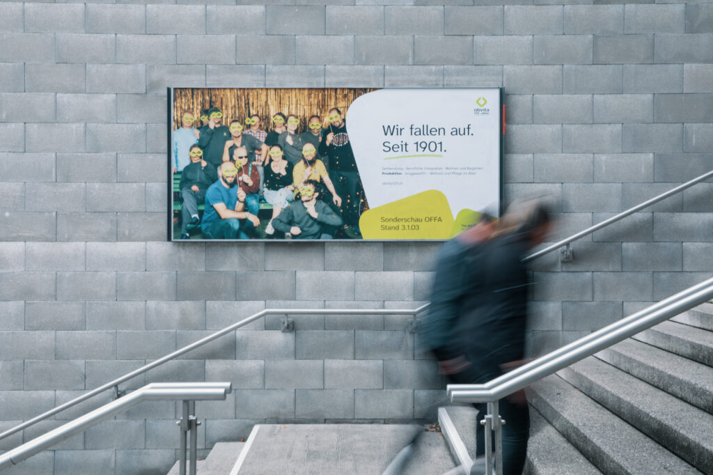 Eine verschwommene Person geht eine Treppe hinauf, vorbei an einer grauen Backsteinwand mit einem großen Plakat, das eine Gruppe lächelnder Menschen zeigt. Auf dem Plakat steht: "Wir fallen auf. Seit 1901." und enthält zusätzlichen deutschen Text.