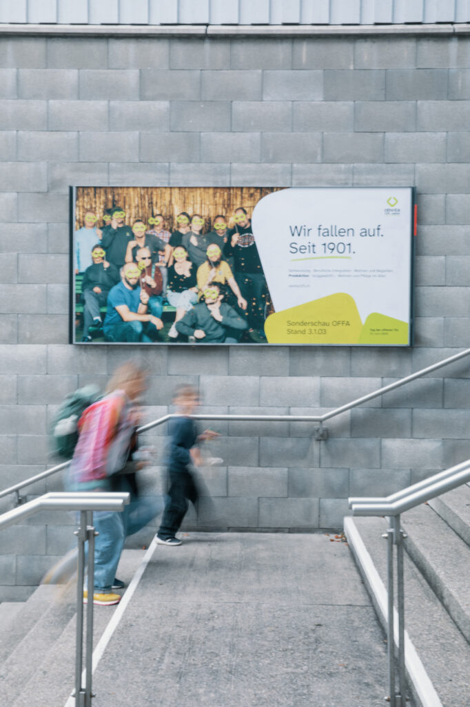 Ein verschwommenes Bild von Menschen, die eine Treppe hinuntergehen, vor einer Wand mit einer großen Werbetafel, die ein Gruppenfoto und den deutschen Text "Wir fallen auf. Seit 1901.".