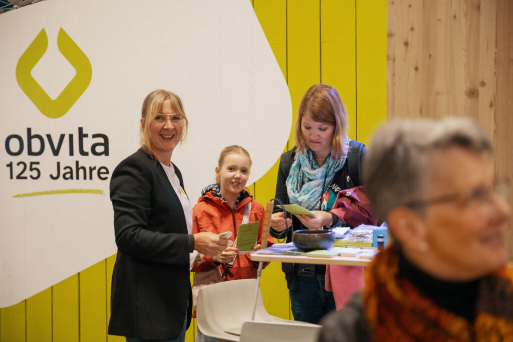Drei Frauen und ein Mädchen stehen bei einer Veranstaltung an einem Tisch mit Flugblättern. Auf einem Schild steht "obvita 125 Jahre". Der Hintergrund ist gelb und holzgetäfelt. Alle lächeln oder sind in ein Gespräch vertieft.