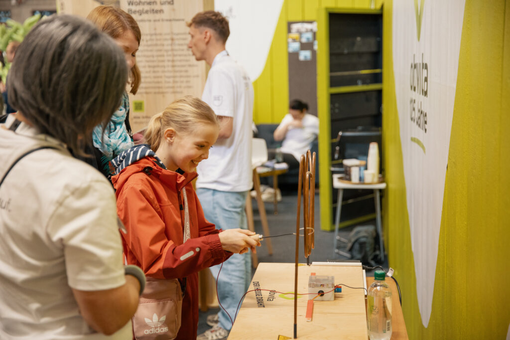 Ein junges Mädchen in einer roten Jacke lächelt, während es sich bei einer Indoor-Veranstaltung mit einer pädagogischen Holzaktivität beschäftigt. Zwei Erwachsene stehen in der Nähe, und andere Personen sind im Hintergrund zu sehen.
