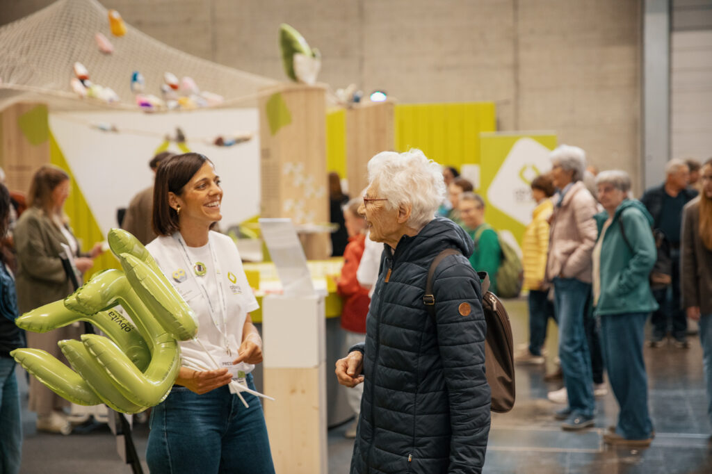 Eine junge Frau, die grüne aufblasbare Objekte in der Hand hält, lächelt und unterhält sich mit einer älteren Frau in einer belebten Indoor-Veranstaltung mit Auslagen und anderen Menschen im Hintergrund.
