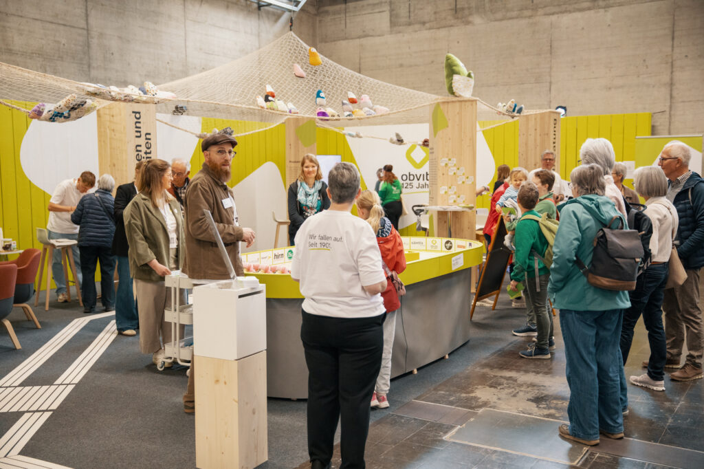 Eine Gruppe von Menschen versammelt sich um einen interaktiven Ausstellungsstand mit hölzernen Strukturen und spielerischen Dekorationen, darunter ein Netz mit Schuhen und Spielzeug darüber. Die Atmosphäre ist lebhaft, und es scheint sich um einen Innenraum zu handeln.
