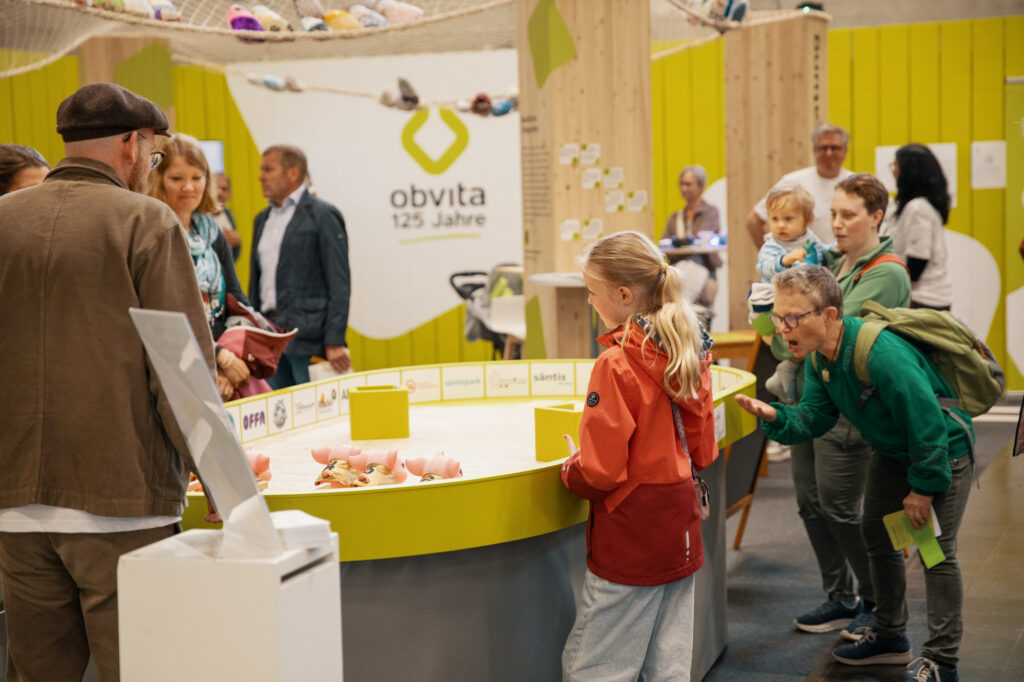 Eine Gruppe von Menschen, darunter Erwachsene und Kinder, versammelt sich bei einer Indoor-Veranstaltung um einen gelben ovalen Ausstellungstisch mit Spielzeugtieren. Im Hintergrund ist ein grünes "obvita 125 Jahre"-Schild zu sehen.