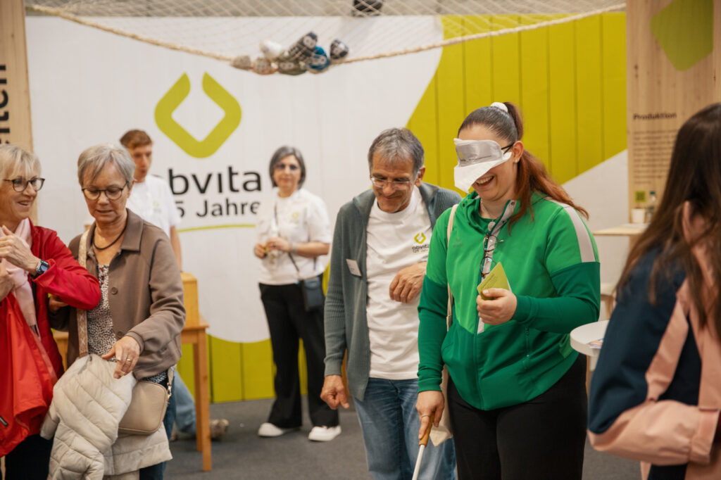 Eine Gruppe von Menschen in einem Haus, einige lächelnd, eine Frau in einer grünen Jacke mit verbundenen Augen und einem Stock in der Hand, die von einer anderen Person geführt wird. An der Wand im Hintergrund sind ein Logo und ein Text zu sehen.