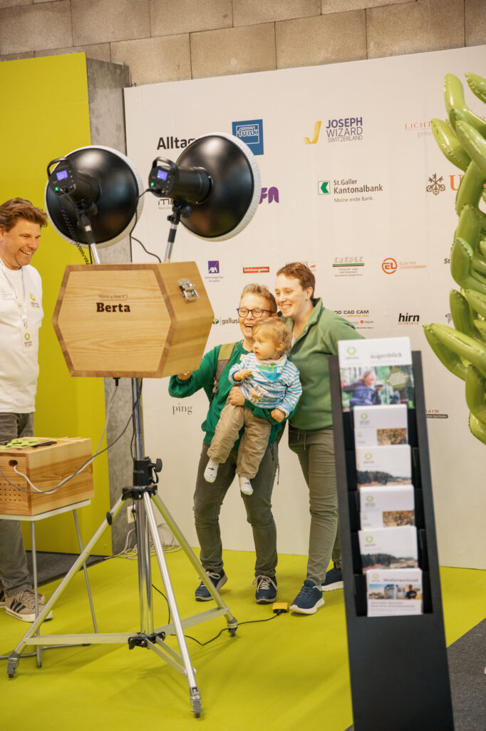 Zwei Frauen und ein kleines Kind lächeln und posieren für ein Foto vor einem Fotoautomaten bei einer Indoor-Veranstaltung. Hinter ihnen befindet sich eine Kulisse mit verschiedenen Sponsorenlogos und einer grünen Anzeigetafel.