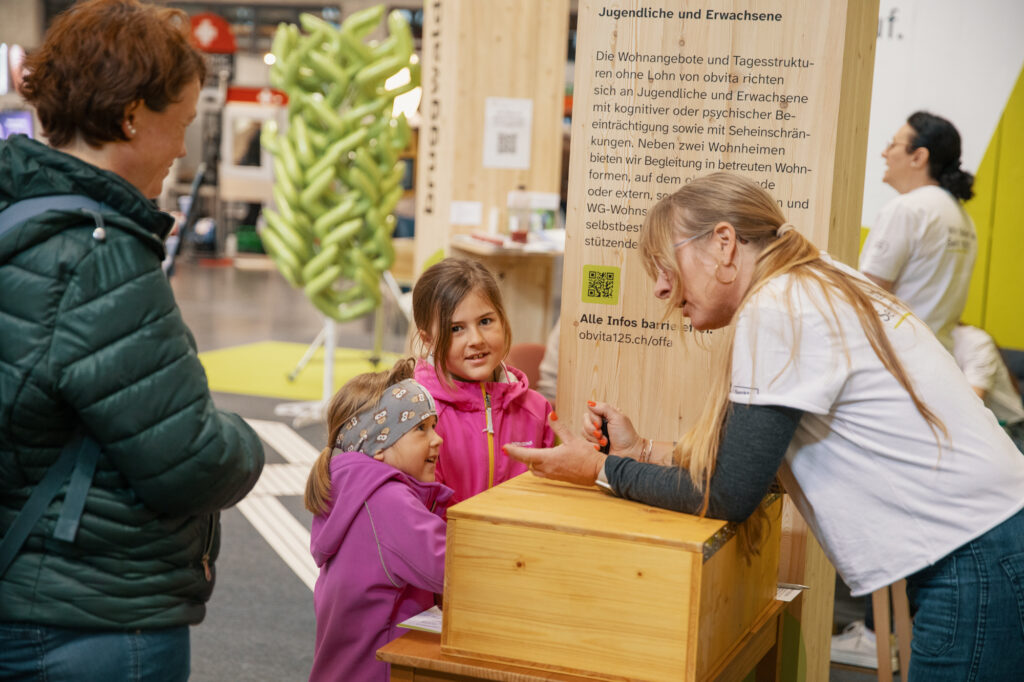 Eine Frau unterhält sich mit zwei lächelnden jungen Mädchen an einem überdachten Informationsstand. Eine andere Frau steht in der Nähe, und im Hintergrund ist eine Person in Weiß zu sehen. Informationsschilder und Holzkonstruktionen sind vorhanden.