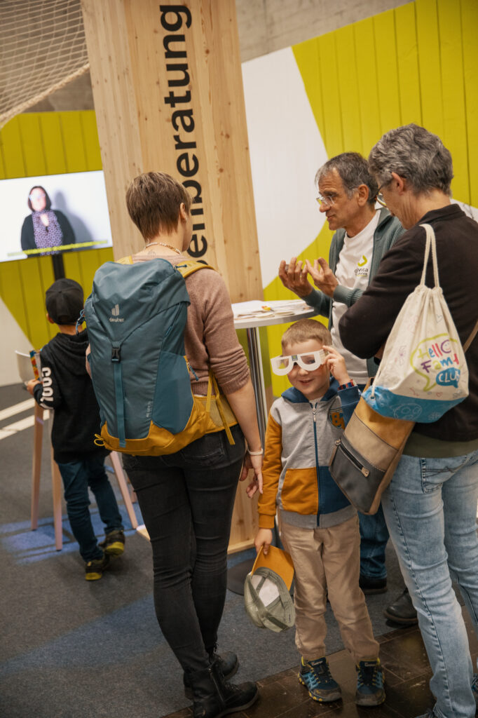 Eine Gruppe von Erwachsenen und Kindern unterhält sich bei einer Indoor-Veranstaltung in der Nähe einer Holzsäule mit der Aufschrift "Beratung". Ein Kind trägt eine Papierbrille, während auf einem Fernsehbildschirm im Hintergrund das Gesicht einer Frau zu sehen ist.
