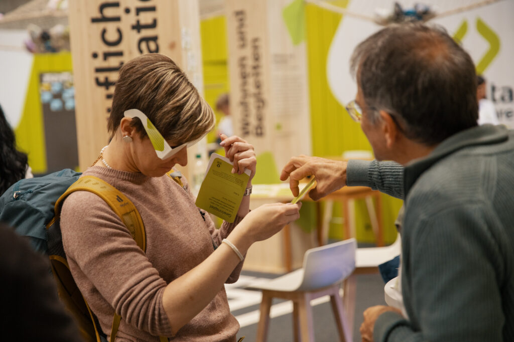 Eine Frau, die eine Virtual-Reality-Brille trägt, interagiert mit einer kleinen Karte, während ein älterer Mann neben ihr auf einer Indoor-Veranstaltung oder -Ausstellung mit grünem und hölzernem Dekor gestikuliert.