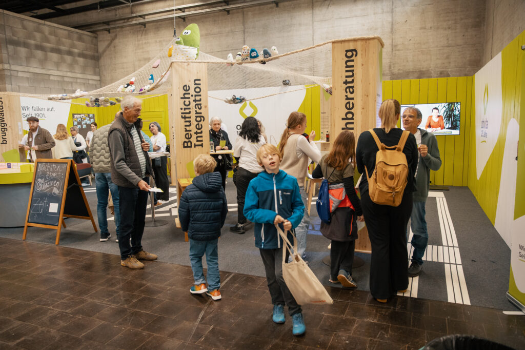 Menschen verschiedenen Alters interagieren an einem Veranstaltungsstand mit Schildern zur beruflichen Integration und Beratung. Kinder und Erwachsene sind mit Aktivitäten beschäftigt; im Hintergrund sind Informationsdisplays und Dekorationen zu sehen.