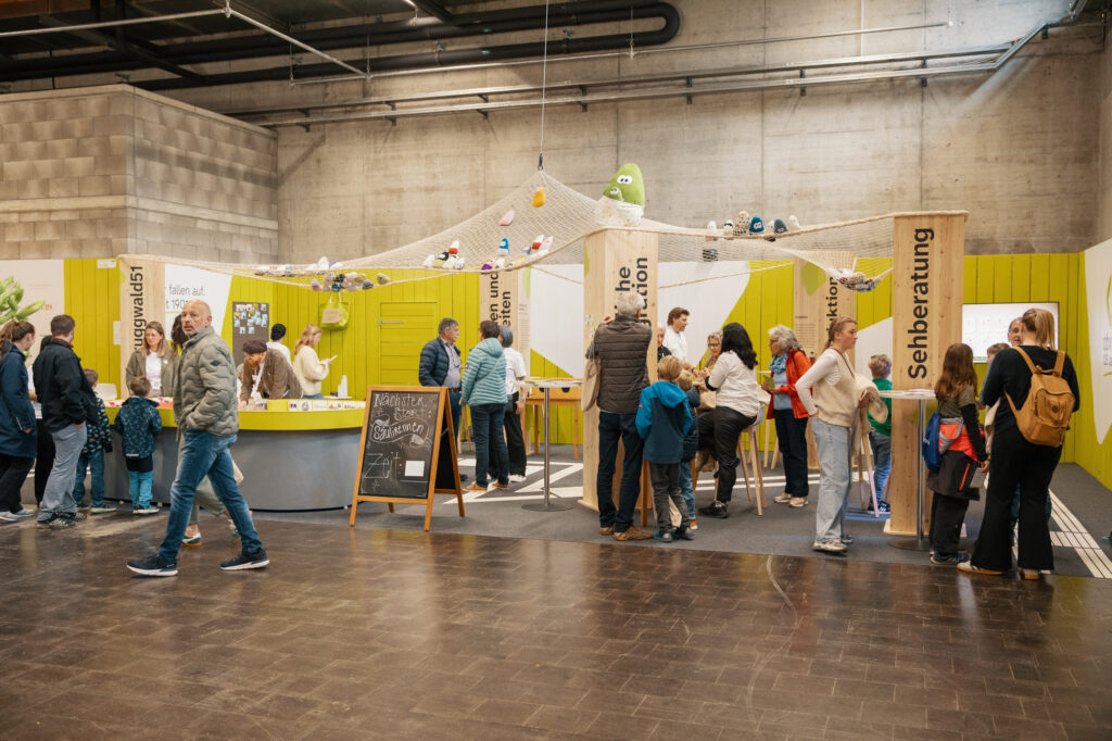 Eine Gruppe von Menschen interagiert an verschiedenen Ständen in einer hell dekorierten Ausstellungshalle mit grünen Wänden und verspielten Dekorationen. Einige lesen Auslagen, andere unterhalten sich oder warten in Schlangen.