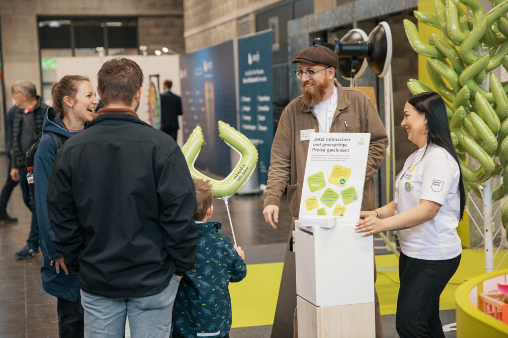 Eine Gruppe von Erwachsenen und ein Kind unterhalten sich lächelnd mit zwei Ausstellern an einem Stand. Eine Frau verteilt grüne Luftballonformen. Im Vordergrund sind ein Schild und ein Informationsstand zu sehen. Der Schauplatz scheint eine Indoor-Veranstaltung oder eine Messe zu sein.