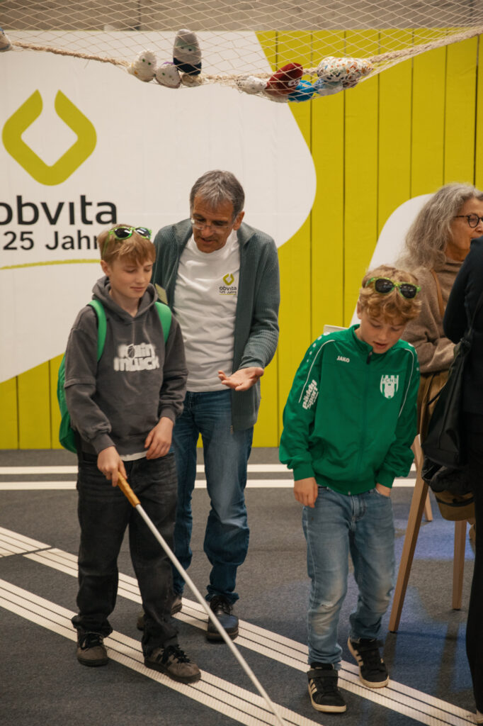 Ein Mann führt zwei Jungen, von denen einer einen weißen Stock benutzt, in einem Innenraum mit leuchtend gelben Wänden und einem Netz darüber. Die Szene scheint pädagogisch oder interaktiv zu sein, mit Menschen, die sich mit einer Aktivität beschäftigen.