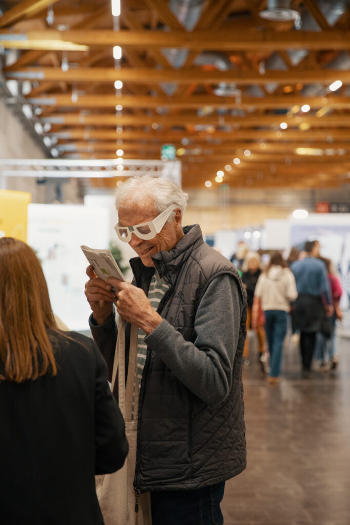 Ein älterer Mann mit einer weißen Papierbrille und einer grauen Weste liest eine Broschüre, während er in einem Innenraum auf einer belebten Veranstaltung oder Ausstellung mit Menschen und hölzernen Deckenbalken im Hintergrund steht.