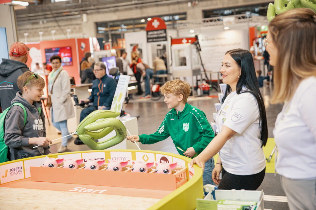 Zwei Frauen und zwei Jungen spielen ein Spiel bei einer Indoor-Veranstaltung mit verschiedenen Ständen im Hintergrund. Eine Frau und ein Junge halten aufblasbare grüne Ringe, und alle scheinen zu lächeln und beschäftigt zu sein.