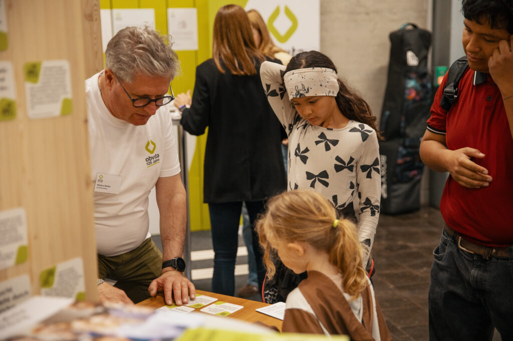 Ein Mann in einem weißen Hemd erklärt zwei jungen Mädchen an einem Tisch während einer Indoor-Veranstaltung etwas; eine Frau und ein weiterer Mann stehen in der Nähe und beobachten. Auf dem Tisch und an den Wänden ist Informationsmaterial ausgelegt.