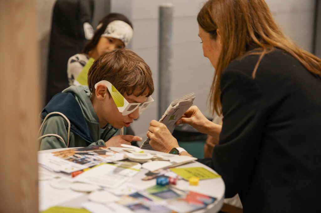Ein Junge mit einer weißen Brille beugt sich über einen Tisch und untersucht ein Tablet, das von einer erwachsenen Frau gehalten wird. Auf dem Tisch liegen Papiere, Broschüren und kleine Gegenstände verstreut. Ein weiteres Kind ist im Hintergrund zu sehen.