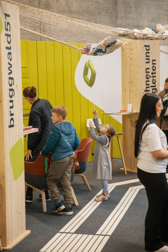 Ein junges Mädchen greift nach einem grünen Luftballon, während ein Junge und ein Erwachsener in der Nähe in einer Innenausstellung mit Holzstrukturen und gelb-grünen Akzenten stehen. Im Hintergrund sind weitere Personen zu sehen.