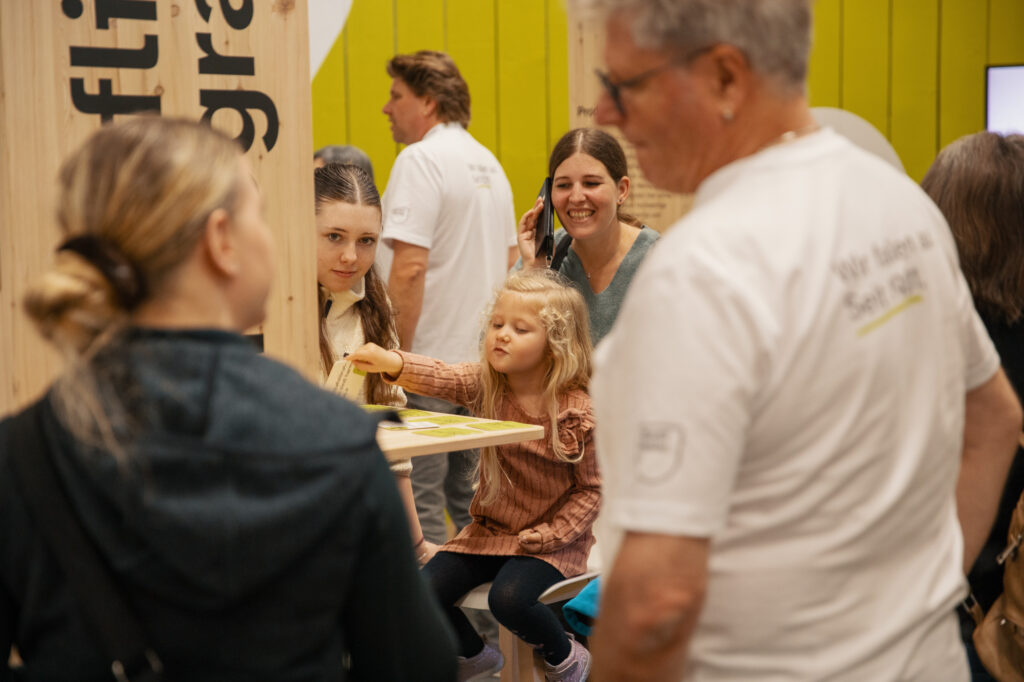 Eine Gruppe von Menschen, darunter Kinder und Erwachsene, versammelt sich bei einer Indoor-Veranstaltung um einen Holztisch. Ein junges Mädchen in einem rosa Kleid greift nach einem Gegenstand auf dem Tisch, während andere zuschauen und sich austauschen.