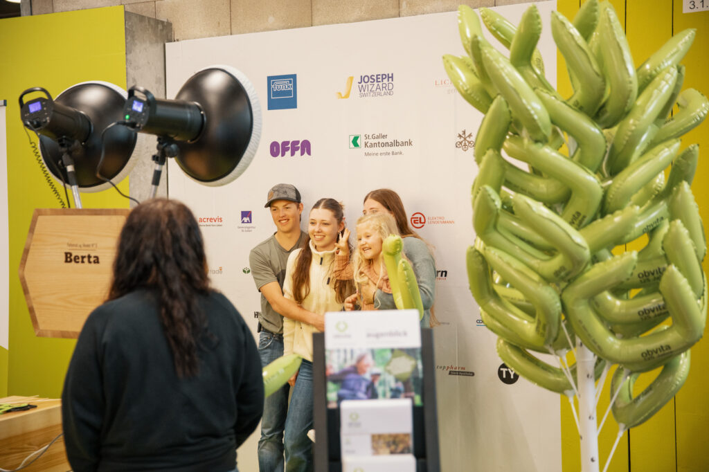 Eine Gruppe von vier Personen posiert für ein Foto vor einer Kulisse mit verschiedenen Logos, während eine Frau eine Kameraausrüstung bedient. Auf der rechten Seite des Bildes sind grüne Luftballondekorationen zu sehen.