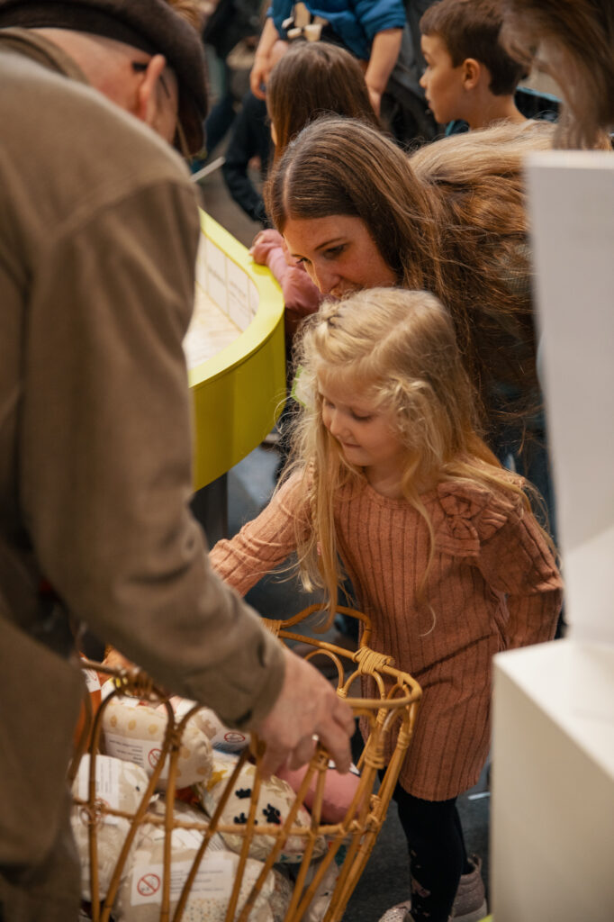 Eine Frau und ein junges Mädchen mit langen blonden Haaren betrachten Gegenstände in einem Weidenkorb, der von einem Mann gehalten wird. Im Hintergrund sind andere Kinder und Erwachsene in einem belebten Innenbereich zu sehen.