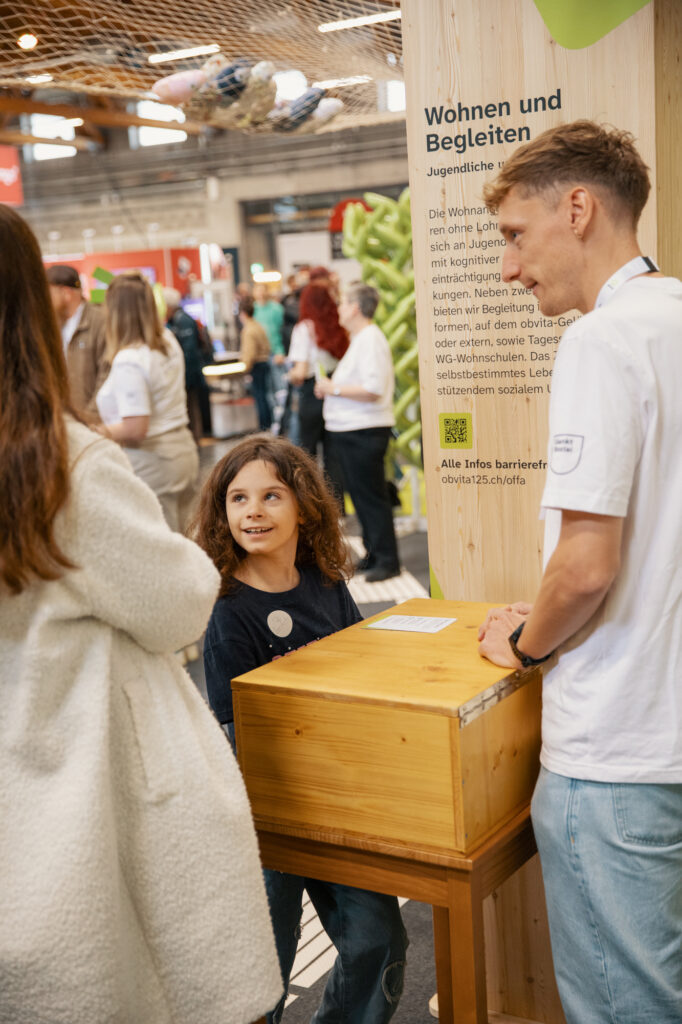 Ein lächelndes Kind mit langen braunen Haaren steht an einer Holzkiste und blickt zu einer Frau und einem Mann in weißen Hemden bei einer Indoor-Veranstaltung auf. Im Hintergrund sind Menschen und Informationsdisplays zu sehen.
