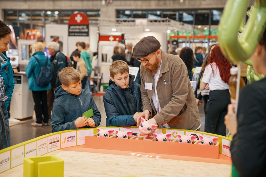 Drei Jungen und ein Mann mit Bart und Mütze stehen bei einer Indoor-Veranstaltung um einen runden Tisch herum und begutachten rosa Spielzeugtiere, die an einer Startlinie aufgereiht sind. Im Hintergrund sind Menschen und Stände zu sehen.