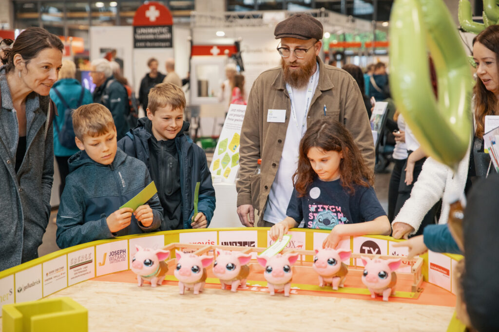 Eine Gruppe von Erwachsenen und Kindern versammelt sich bei einer Indoor-Veranstaltung oder -Ausstellung um eine sandkastenähnliche Ausstellung mit mehreren rosa Spielzeug-Schweinerobotern. Die Kinder wirken engagiert und neugierig, während sie mit der Ausstellung interagieren.