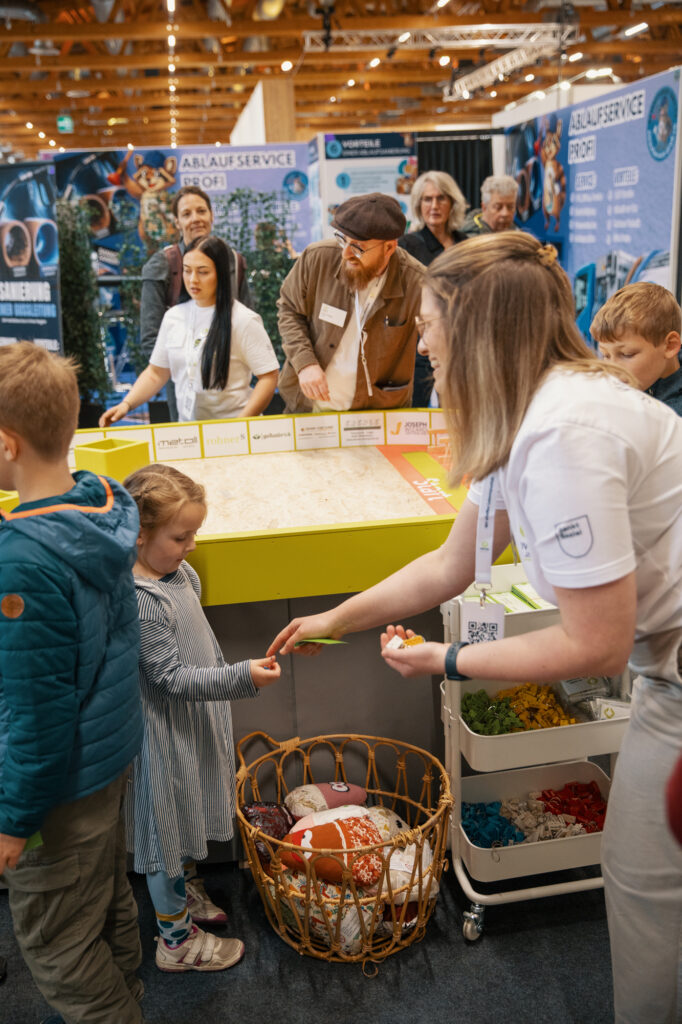Eine Frau überreicht einem jungen Mädchen an einem bunten Stand, umgeben von Kindern und Erwachsenen in einer belebten Veranstaltungshalle, einen kleinen Gegenstand. Im Hintergrund sind ein Korb mit Spielzeug und Informationsplakate zu sehen.