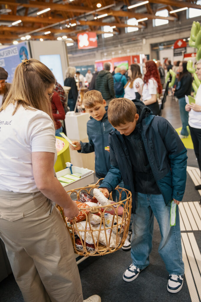 Eine Frau hält einen Korb mit verschiedenen Gegenständen in der Hand, während zwei Jungen in Jacken darin herumstöbern und Gegenstände bei einer geschäftigen Indoor-Veranstaltung mit anderen Menschen und Ständen im Hintergrund heraussuchen.
