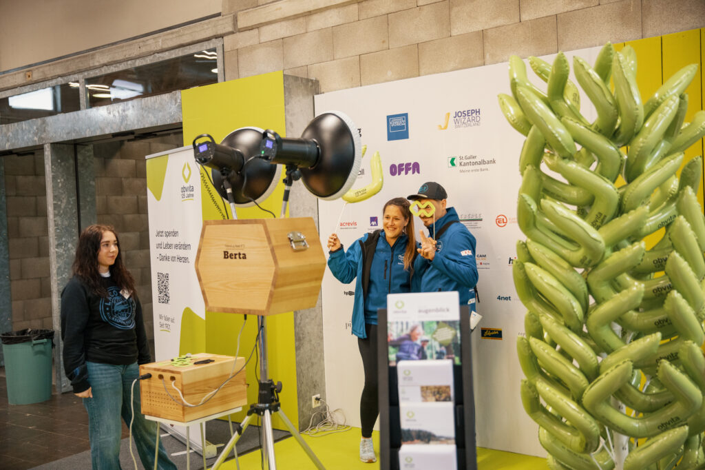 Zwei Personen posieren für ein Foto vor einem Markenhintergrund, sie tragen blaue Jacken und halten grüne Requisiten. Eine Frau steht in der Nähe. Im Vordergrund befinden sich ein hölzerner Fotoautomat und grüne aufblasbare Dekorationen.