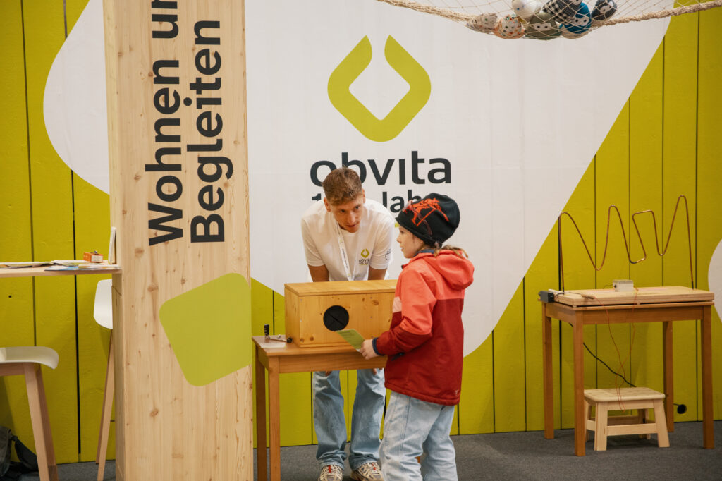 Ein junger Erwachsener in einem weißen Hemd hilft einem Kind in einer roten Jacke an einem Ausstellungsstück mit Holzkonstruktionen. Im Hintergrund ist eine gelbe Wand mit dem "obvita"-Logo und Text in deutscher Sprache zu sehen.
