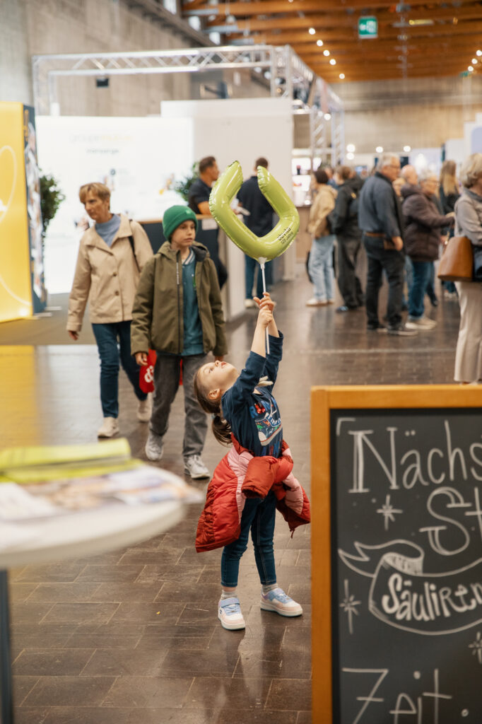 Ein kleines Kind in einer belebten Veranstaltungshalle balanciert einen grünen Luftballon auf einem Stock, während es nach oben schaut, umgeben von Menschen, die spazieren gehen, und einem Kreideschild im Vordergrund.