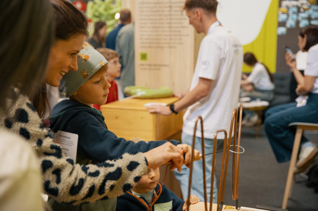Eine Frau und zwei kleine Kinder interagieren mit einem wissenschaftlichen Exponat zum Anfassen. Im Hintergrund des belebten Veranstaltungsraums sind weitere Personen mit Aktivitäten beschäftigt.