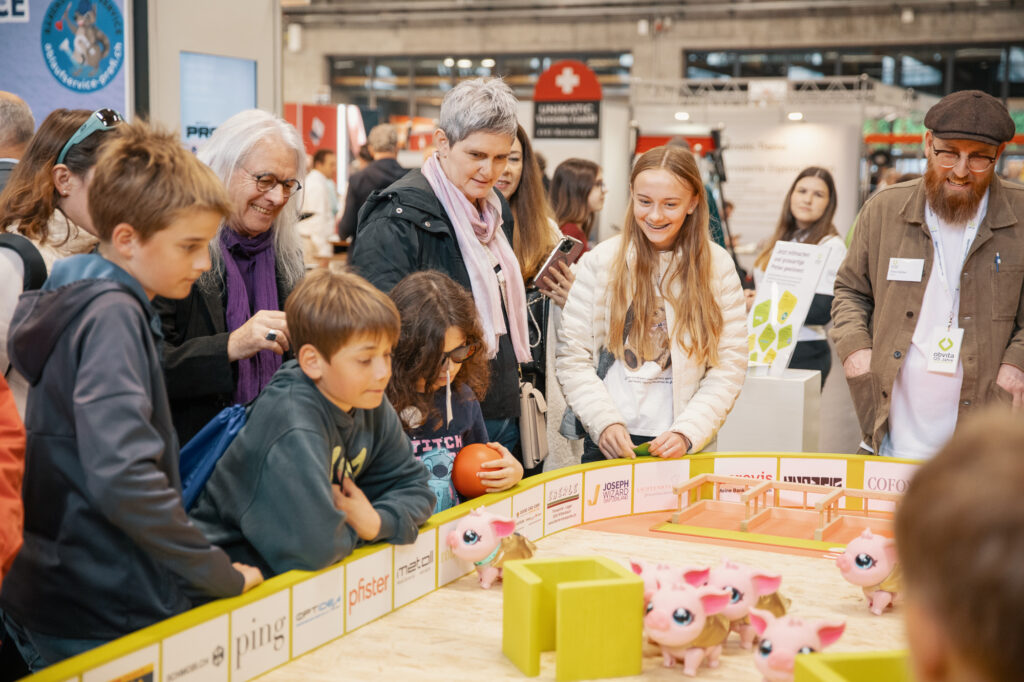 Eine Gruppe von Kindern und Erwachsenen versammelt sich um einen Tisch und beobachtet rosa Spielzeugschweine in einer Spielarena bei einer Indoor-Veranstaltung oder Ausstellung. Die Leute lächeln und sind mit der Aktivität beschäftigt.