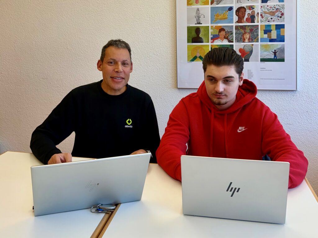 Zwei Männer sitzen nebeneinander an einem Tisch und arbeiten jeweils an einem HP-Laptop. Einer trägt ein schwarzes Hemd, der andere einen roten Kapuzenpulli. Sie befinden sich in einem hellen Raum mit einem bunten Poster an der Wand hinter ihnen.