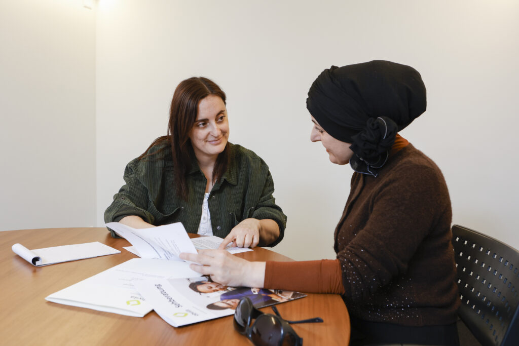 Zwei Frauen sitzen an einem runden Tisch, unterhalten sich und sehen sich Dokumente an. Eine Frau trägt eine grüne Jacke, die andere einen braunen Pullover und ein schwarzes Kopftuch. Auf dem Tisch vor ihnen sind Papiere und eine Broschüre ausgebreitet.
