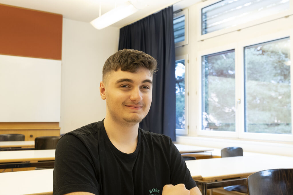 Ein junger Mann mit kurzen Haaren und einem leichten Lächeln sitzt in einem hellen Klassenzimmer mit leeren Tischen, großen Fenstern und einem Whiteboard im Hintergrund.