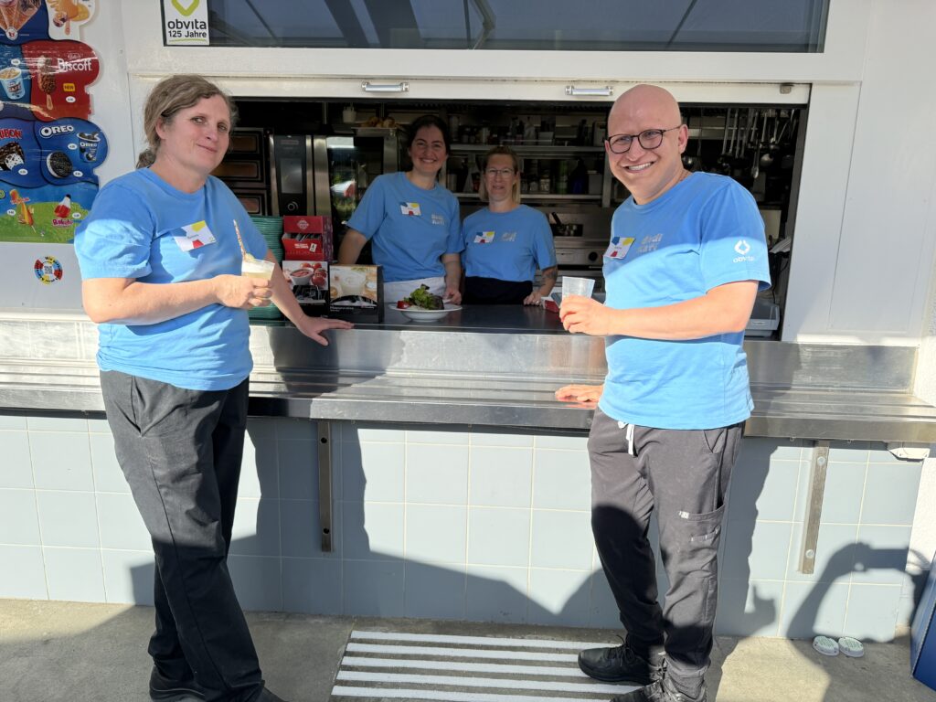 Vier Personen in blauen Hemden stehen lächelnd an einem Tresen vor einem Imbissstand oder Café und halten Eis und Getränke in der Hand. Zwei von ihnen befinden sich im Inneren des Standes, zwei im Freien. Es ist sonnig und hell.