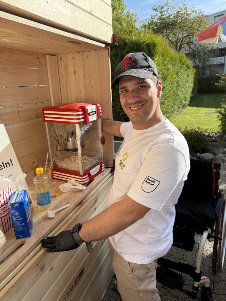 Ein lächelnder Mann in weißem T-Shirt und schwarzer Mütze bereitet an einem sonnigen Tag an einem Holzstand im Freien Popcorn mit einer rot-weißen Popcornmaschine zu. Auf der Theke liegen verschiedene Popcorn-Zubehörteile.
