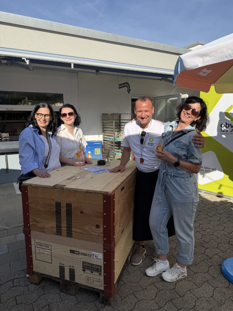 Vier lächelnde Erwachsene stehen im Freien um eine Holzkiste herum und halten Getränke in der Hand. Zwei Frauen tragen Sonnenbrillen und helle Kleidung; ein Mann in einem weißen Hemd steht zwischen ihnen. Die Umgebung wirkt zwanglos und sonnig, Essen und Trinken sind in der Nähe.