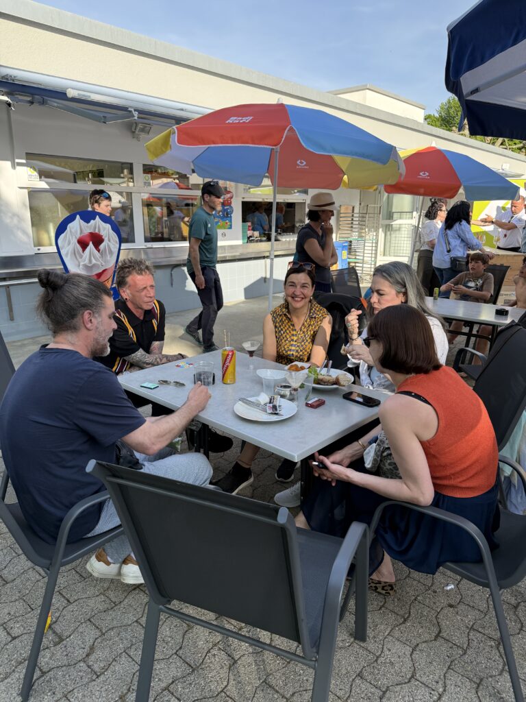 Vier Personen sitzen an einem Tisch im Freien, essen und unterhalten sich unter bunten Sonnenschirmen. Andere Menschen stehen und gehen im Hintergrund. Leere Teller und Getränke sind auf dem Tisch zu sehen. Die Atmosphäre ist locker und sonnig.