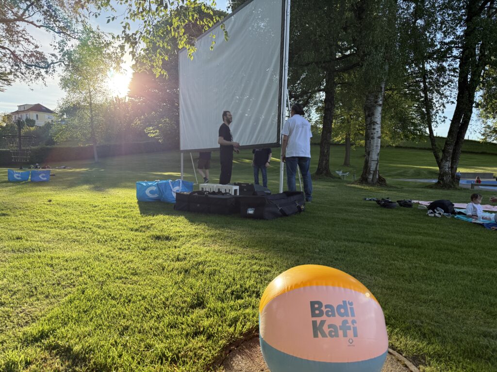 Eine Gruppe von Menschen stellt bei Sonnenuntergang auf einer Wiese eine große Leinwand auf. Im Vordergrund ist ein bunter Strandball mit dem Aufdruck "Badi Kafi" zu sehen. Bäume und verstreutes Veranstaltungsequipment sind zu sehen.
