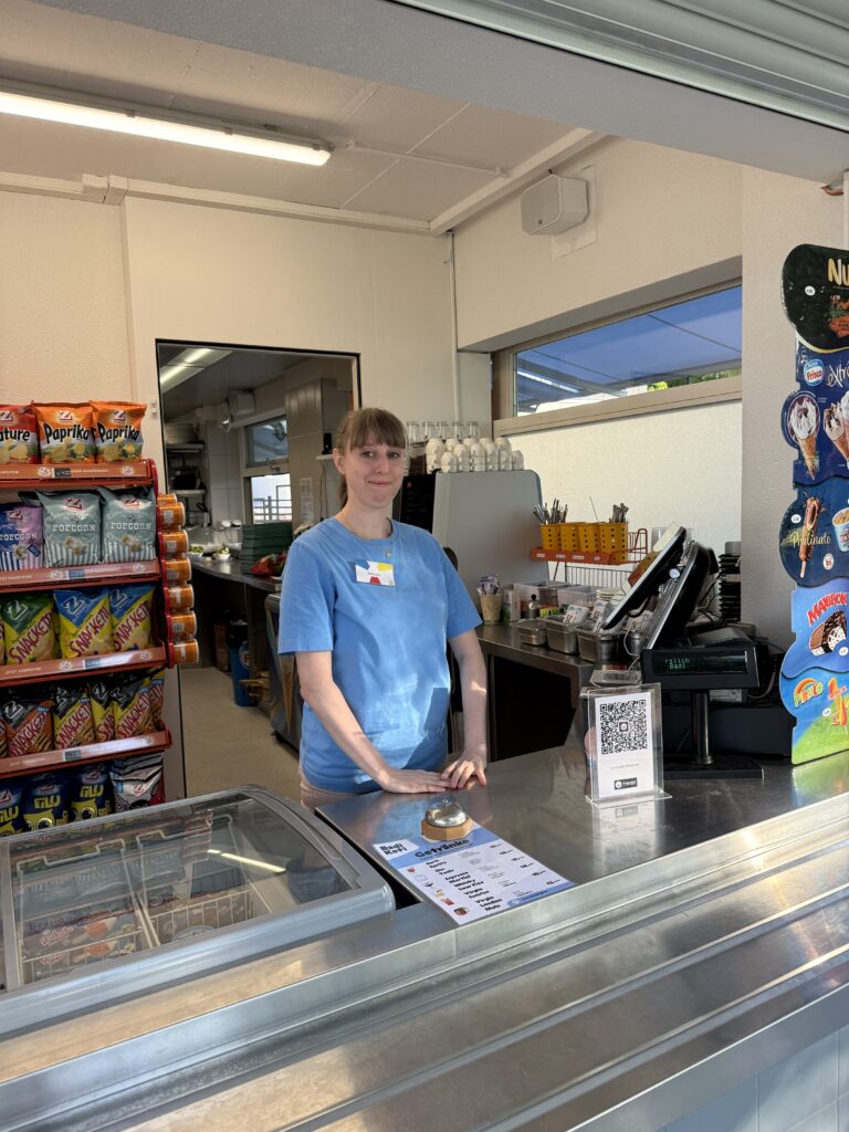 Hinter dem Tresen eines Imbisses steht eine Person mit blauem Hemd und Namensschild. Es gibt Regale mit Chips, eine Registrierkasse, einen QR-Code und ein Fenster, das Sonnenlicht hereinlässt.
