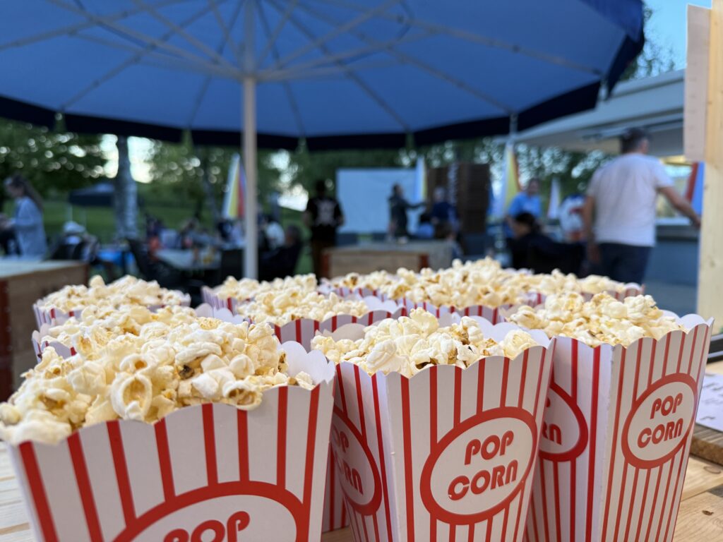 Im Vordergrund stehen mehrere rot-weiß gestreifte Popcornbehälter, die mit Popcorn gefüllt sind. Im Hintergrund versammeln sich Menschen im Freien unter einem großen blauen Regenschirm, was auf eine Veranstaltung oder einen Filmabend im Freien hindeutet.