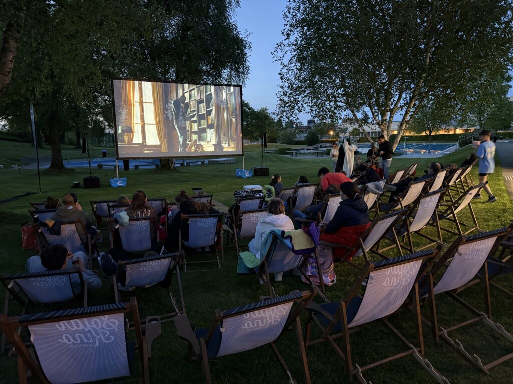 Menschen sitzen auf Liegestühlen auf einer Wiese und sehen sich in der Abenddämmerung einen Film auf einer großen Leinwand an. Bäume umgeben das Gelände, und der Himmel ist tiefblau, wenn der Abend beginnt.