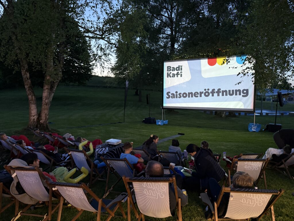 In der Abenddämmerung sitzen die Menschen auf Liegestühlen auf einer Wiese und schauen auf eine große Leinwand, auf der "Badi Kafi Saisoneröffnung" zu sehen ist, umgeben von Bäumen und einer ruhigen, entspannten Atmosphäre.
