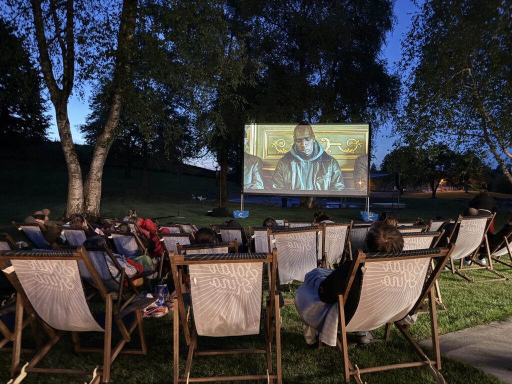 In der Abenddämmerung sitzen Menschen auf Liegestühlen in einem Park und sehen sich einen Film an, der auf eine von Bäumen umgebene Leinwand im Freien projiziert wird. Die Szene auf der Leinwand zeigt einen Mann in einem dunklen Gewand mit einem goldgerahmten Hintergrund.