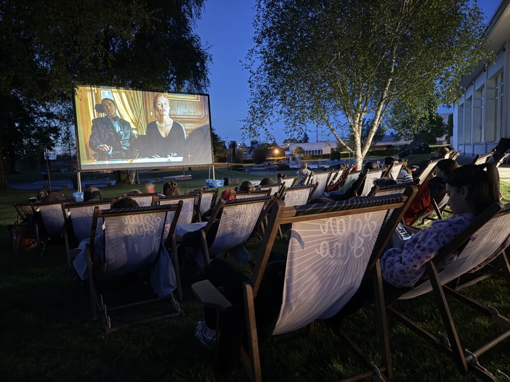 Menschen sitzen nachts auf Liegestühlen im Freien und sehen sich einen Film an, der auf eine große Leinwand projiziert wird. Im Hintergrund sind Bäume, Gras und Gebäude zu sehen, was eine entspannte Open-Air-Kinoatmosphäre schafft.